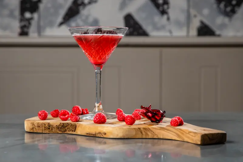 Raspberry hibiscus martini in crystal glass on rustic olive wood board with fresh raspberries garnish