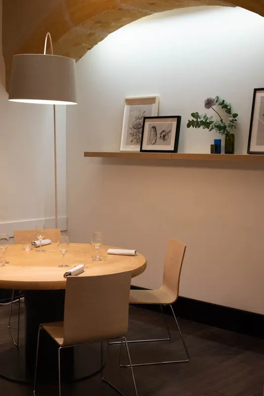 Elegant restaurant dining area with round wooden table, modern chairs, botanical artwork, and arched stone ceiling.