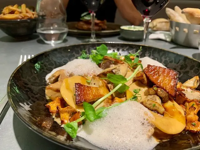 Gourmet wild mushroom dish with crispy bacon, foam sauce, and microgreens in elegant dark bowl at fine dining restaurant