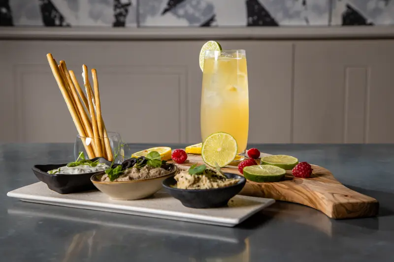 Three Mediterranean dips with breadsticks and a citrus lemonade cocktail garnished with lime on a wooden board.