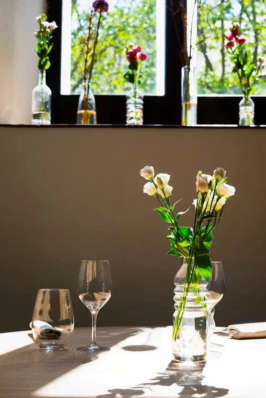 Sunlit restaurant table with white roses in glass vase, wine glasses, and flower-lined windowsill overlooking green trees.