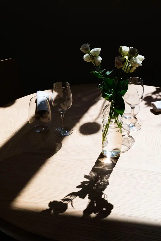 Elegant restaurant table with white roses in glass vase, wine glasses, and dramatic natural lighting on wood surface.