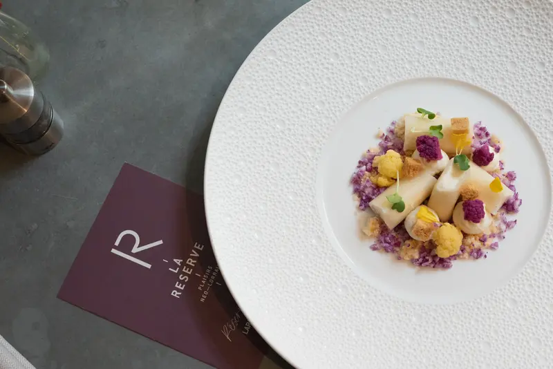 Elegant fine dining dish at La Réserve restaurant featuring white asparagus, purple cauliflower, and edible flowers on textured plate.