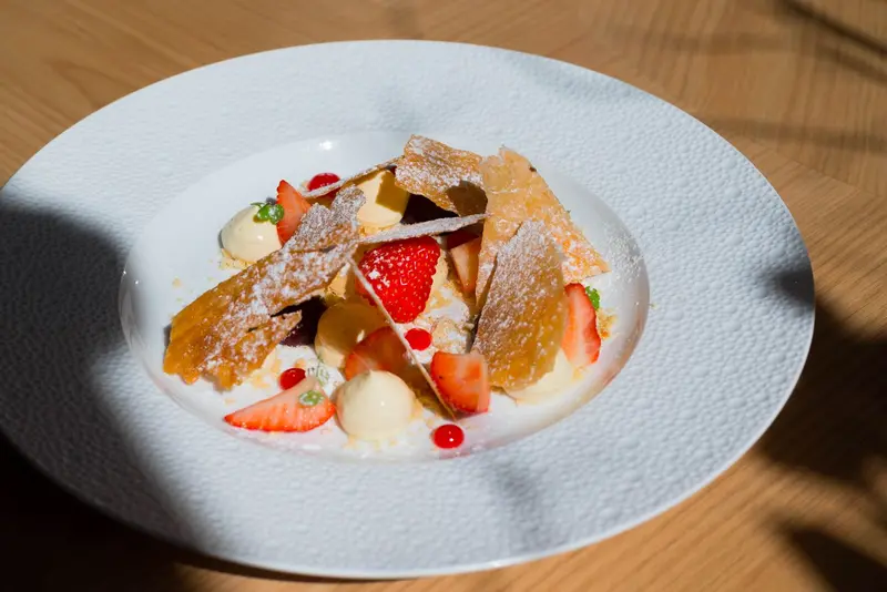 Elegant strawberry dessert with caramelized pastry shards, cream quenelles, and powdered sugar on white plate