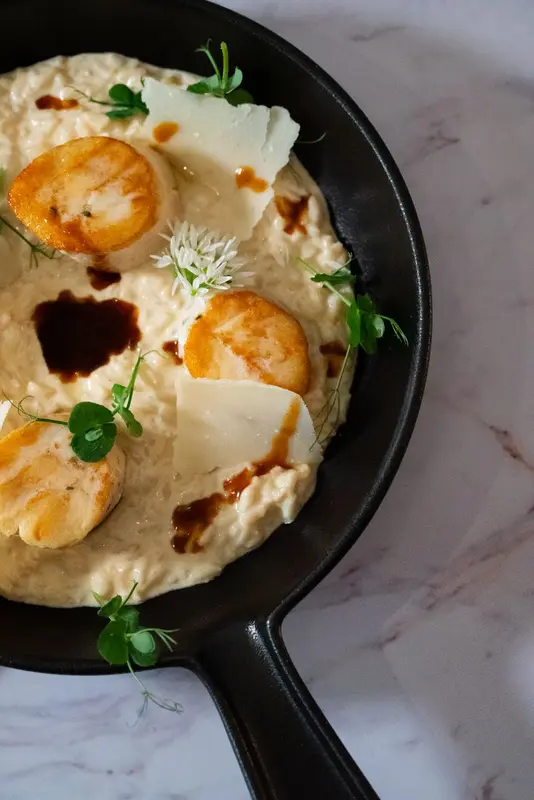 Seared scallops on creamy risotto with shaved parmesan, balsamic drizzle, and microgreens in a cast iron skillet.