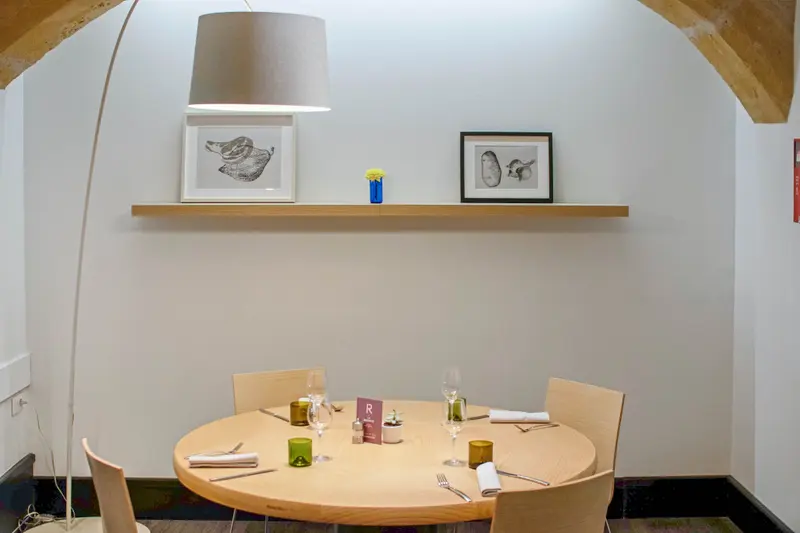 Elegant restaurant dining room with round wooden table set for four, arched stone ceiling, and food artwork on shelf.