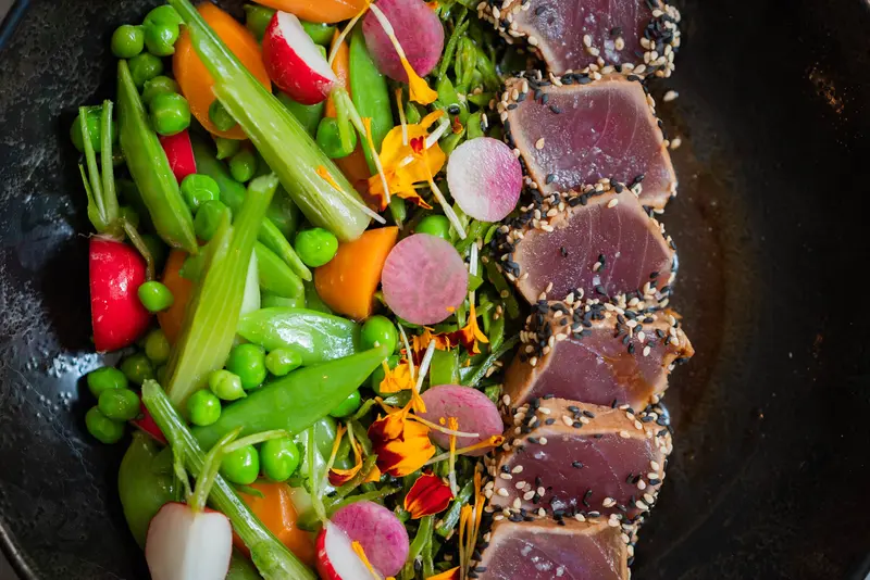 Sesame-crusted seared tuna slices with fresh peas, radishes, snap peas, carrots, and edible flowers on a dark plate.