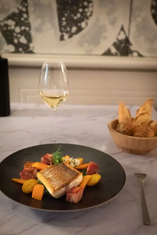 Pan-seared fish fillet with roasted vegetables, bacon, and white wine at an elegant marble-table restaurant setting.