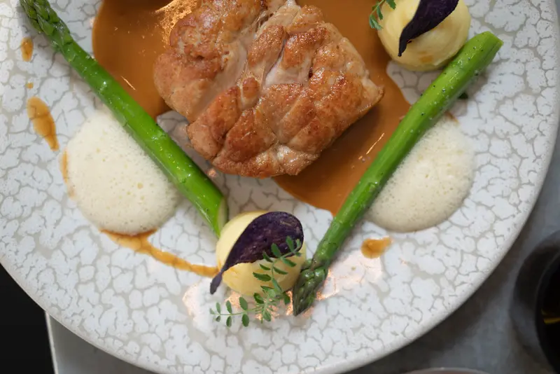 Seared chicken breast with asparagus, rich jus, creamy foam, purple potato chips, and fresh herbs on artisan plate