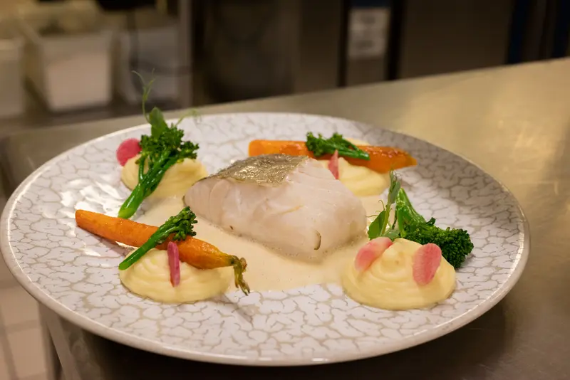 Elegant pan-seared white fish fillet with roasted carrots, broccolini, piped potato purée, and cream sauce on artisan plate.