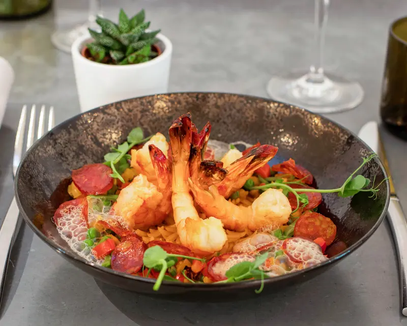 Grilled prawns with chorizo, peas, pasta, and microgreens in a dark bowl at an upscale restaurant