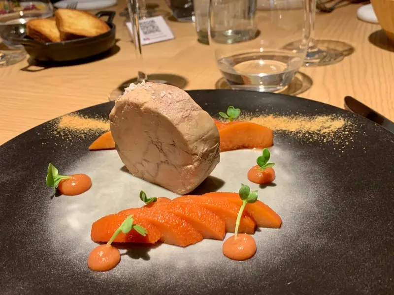 Foie gras terrine with glazed carrots, microgreens, and carrot purée dots on a black plate at a fine dining restaurant.