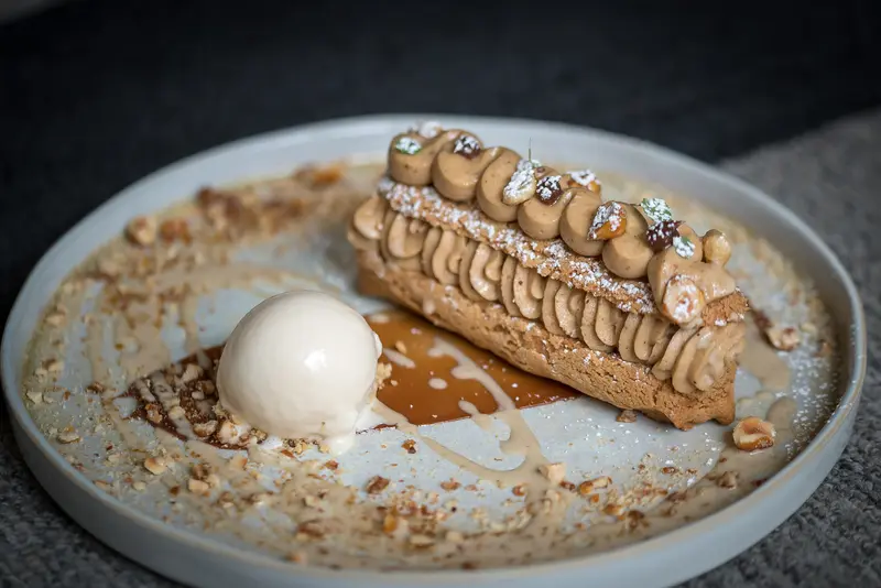 Elegant hazelnut Paris-Brest pastry with caramel sauce and vanilla ice cream scoop, dusted with powdered sugar on ceramic plate.