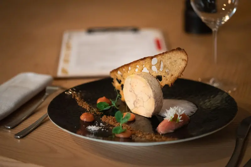 Elegant foie gras terrine with toasted brioche, carrot purée, edible flowers, and spiced crumble on a dark slate plate.