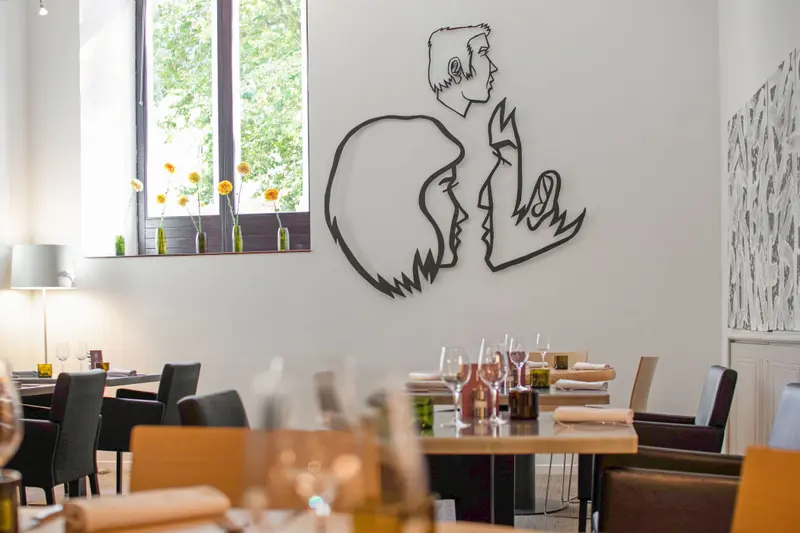 Elegant restaurant dining room with modern line-art wall sculpture, wine glasses, wood tables, and yellow flowers by bright window.
