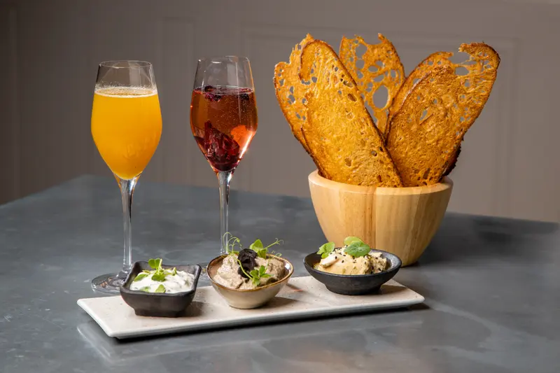 Three dips with crispy toast crostini, a mimosa, and berry spritz cocktail served at an upscale restaurant.