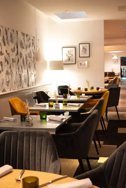 Elegant restaurant dining room with velvet gray and mustard chairs, marble tables, green glassware, and modern artwork on walls.