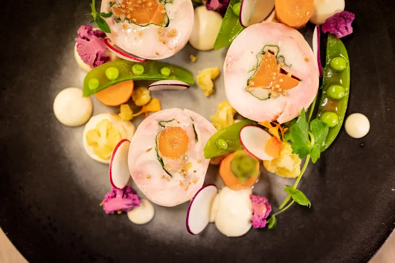 Elegant fine dining dish with pink-wrapped salmon rolls, snap peas, radish, carrots, and purple cauliflower on dark plate