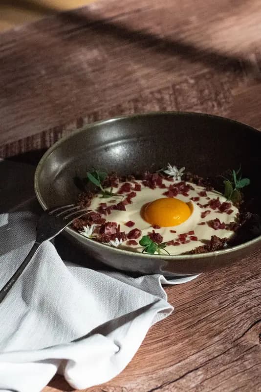 Elegant egg yolk dish with creamy sauce, crispy bits, and microgreens in dark ceramic bowl on rustic wood table