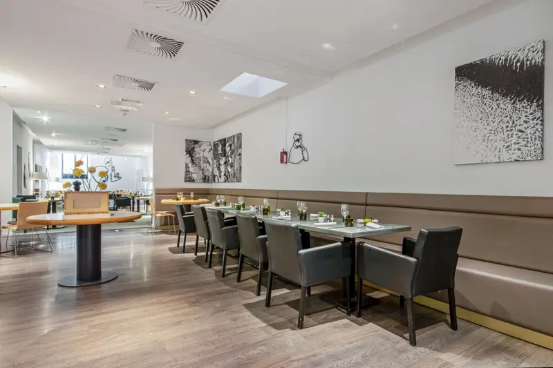 Modern restaurant dining room with banquette seating, gray chairs, set tables, wood floors, and contemporary black-and-white wall art.