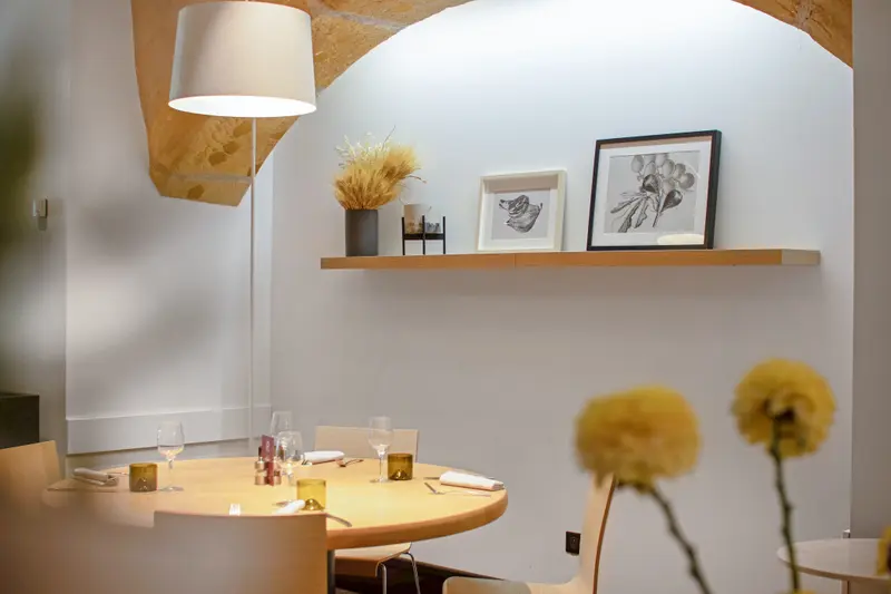 Elegant restaurant dining room with round table set for four, arched stone ceiling, warm lighting, and botanical artwork on shelf.