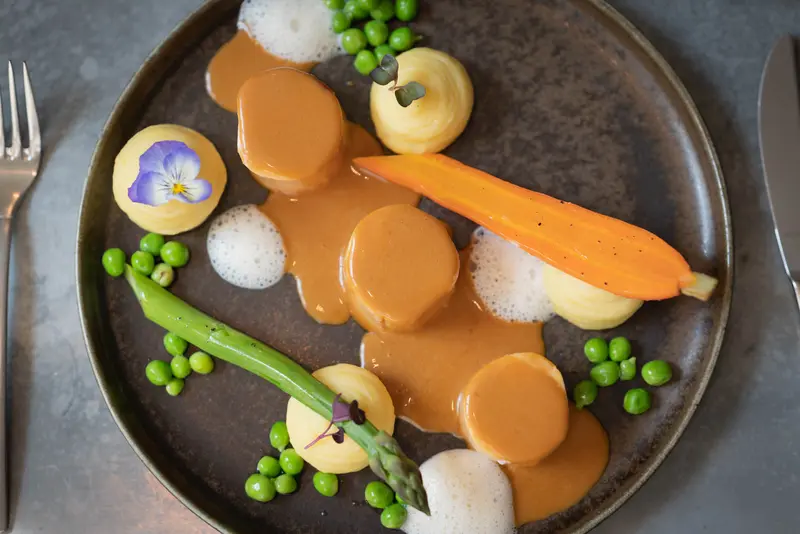 Elegant fine dining plate with duchess potatoes, glazed carrot, asparagus, peas, and rich gravy sauce, garnished with edible pansy flower.