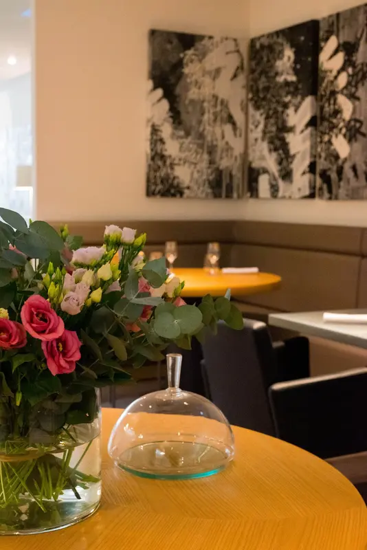Elegant restaurant interior with fresh pink roses, glass cloche, yellow tables, booth seating, and modern black-and-white wall art.