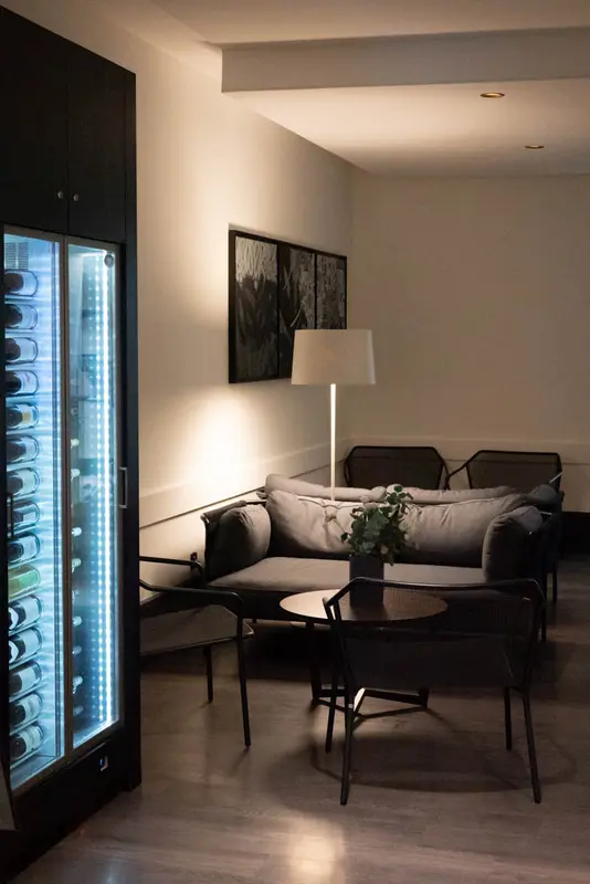 Elegant restaurant lounge with illuminated wine fridge, cozy gray sofa, black chairs, and warm ambient lighting.