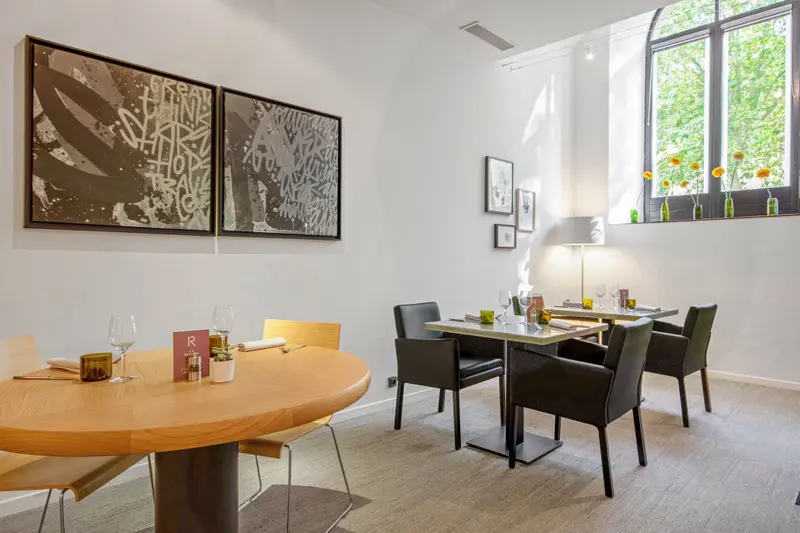 Elegant restaurant dining room with modern art, set tables, wine glasses, and natural light from arched windows.