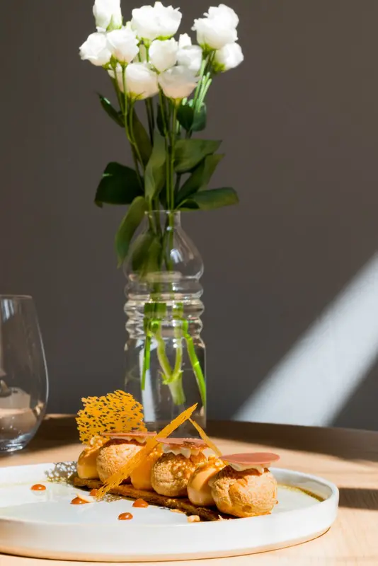 Elegant choux pastry dessert with caramel tuile and sauce on white plate, white roses centerpiece, fine dining restaurant setting.