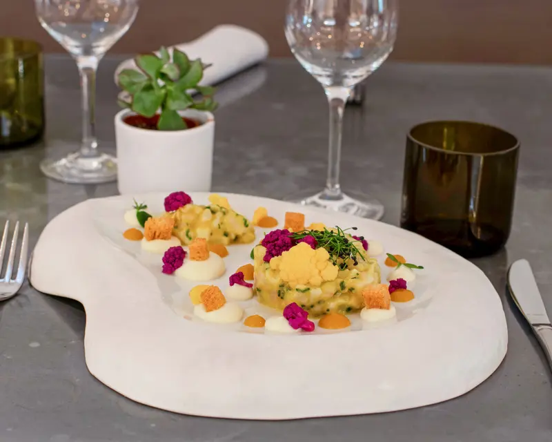 Elegant tartare dish with yellow cauliflower, purple florets, carrot dots, and croutons on a white marble plate at a fine dining restaurant.
