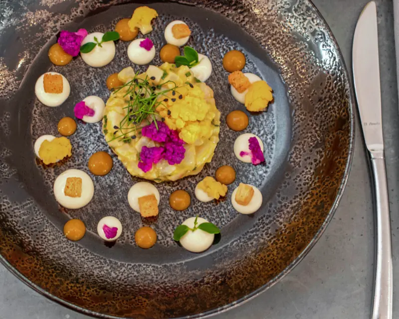 Elegant fine dining dish with colorful cauliflower, cream dots, caramel spheres, and microgreens on dark artisan plate.