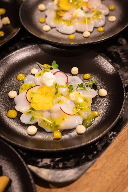 Scallop crudo with yellow edible flowers, radish, herb oil, and cream dots on black plate
