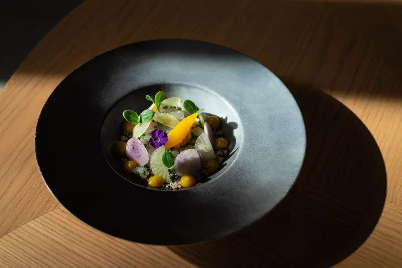 Fine dining dish with quinoa, golden tomatoes, edible flowers, and microgreens on a matte black plate at a modern restaurant.