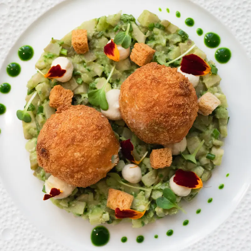 Two golden fried croquettes on pea purée with croutons, cream dots, herb oil, and edible flowers on white plate.