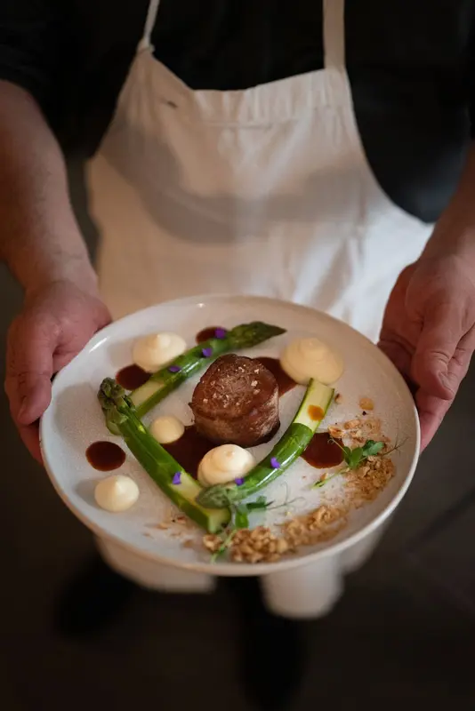 Chef presenting elegant plated dish with asparagus, bacon-wrapped meat medallion, cream dots, and red wine reduction sauce.